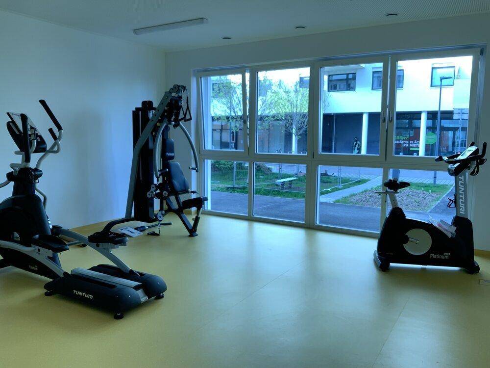 Gemeinschaftlicher Fitnessraum mit verschiedenen Trainingsgeräten und großen Fenstern zum Außenbereich.