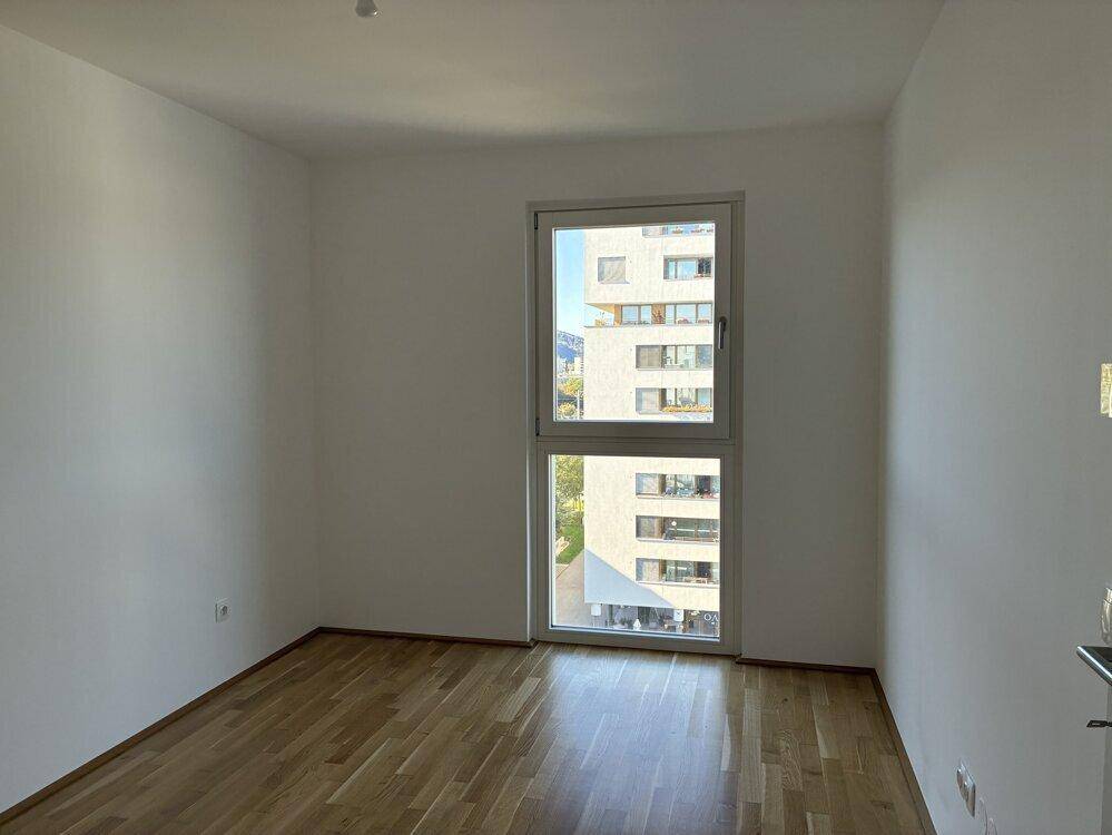 Helles Schlafzimmer mit Parkettboden und großem Fenster, das einen Blick auf ein Nachbargebäude bietet.