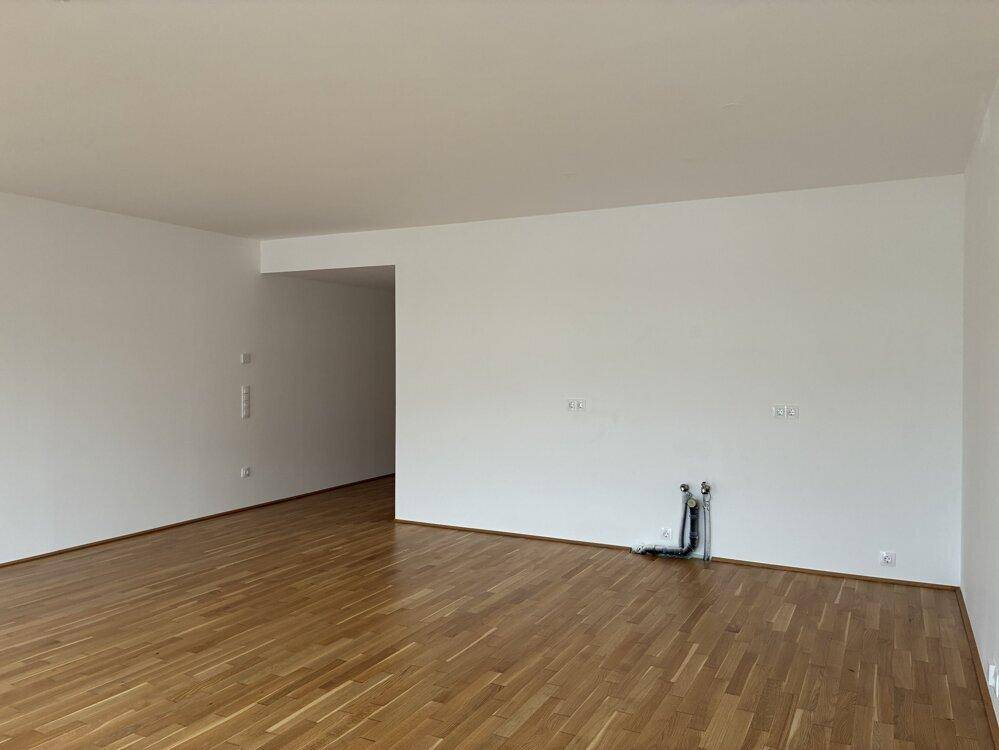 Geräumiger, unmöblierter Wohnbereich mit Parkettboden und Anschlüssen für eine Küche.