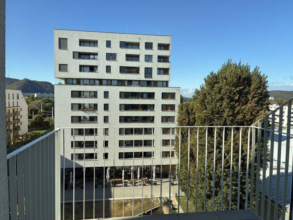 Blick von einem Balkon auf ein gegenüberliegendes modernes Gebäude und die umliegende Berglandschaft.