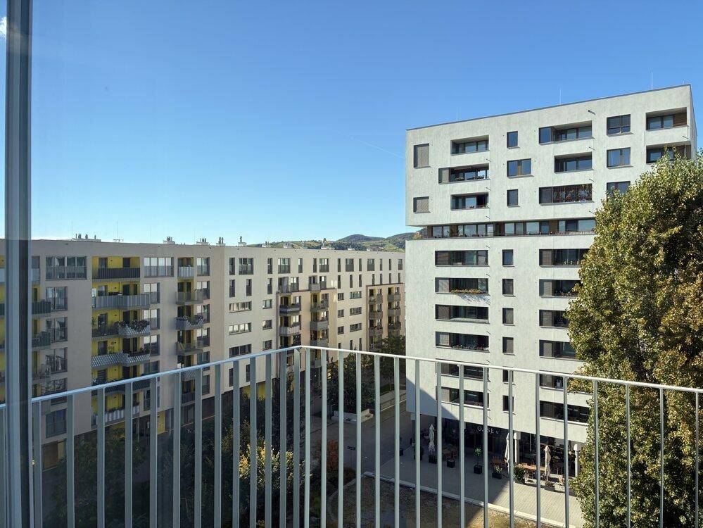 Weitblick vom Balkon auf moderne Gebäude und eine grüne Hügellandschaft unter blauem Himmel.