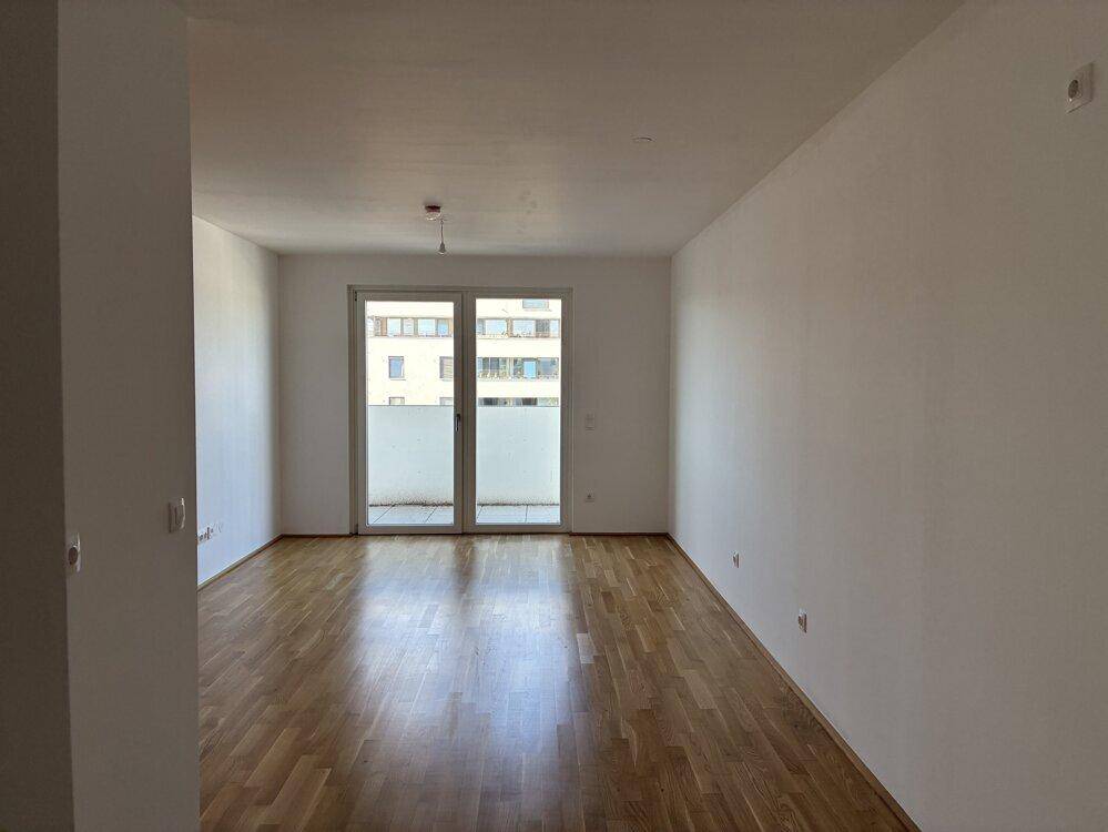 Lichtdurchfluteter Wohnraum mit Holzboden und breiter Balkontür, die den Blick nach draußen öffnet.