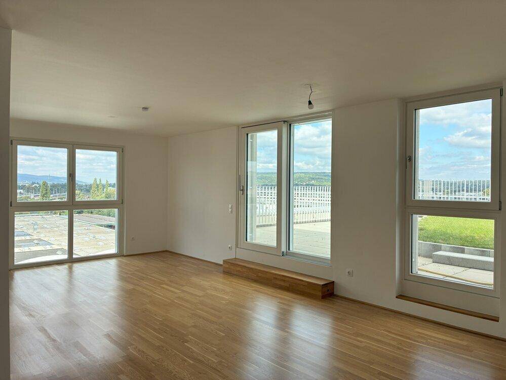 Helles, leeres Wohnzimmer mit Holzboden und großen Fenstern, die Zugang zu einem Außenbereich bieten.