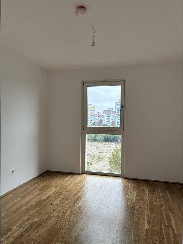 Helles Schlafzimmer mit Parkettboden, weißen Wänden und Fensterblick ins Grüne und auf Gebäude.