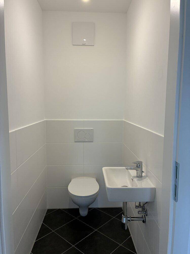 Modernes Gäste-WC mit weißen Fliesen, schwarzem Boden und wandhängendem WC mit kleinem Waschbecken.