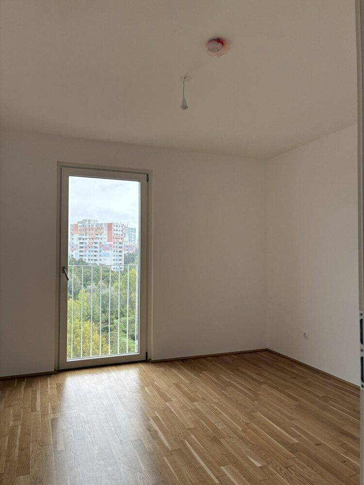 Leeres Schlafzimmer mit Parkettboden, weißen Wänden und Zugang zu einem Balkon mit Stadtblick.