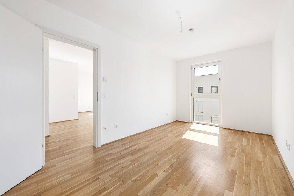 Helles Zimmer mit Holzboden und großem Fenster, verbunden mit einem weiteren Raum.