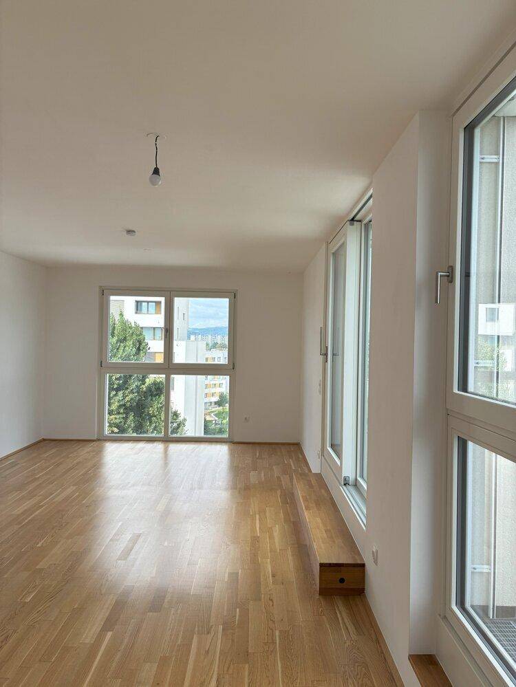 Geräumiges, leeres Zimmer mit Holzboden und großen Fenstern, die einen Blick ins Grüne bieten.