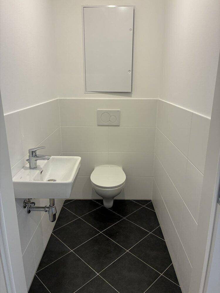 Helles Gäste-WC mit weißen Fliesen, schwarzem Boden und wandhängendem WC.
