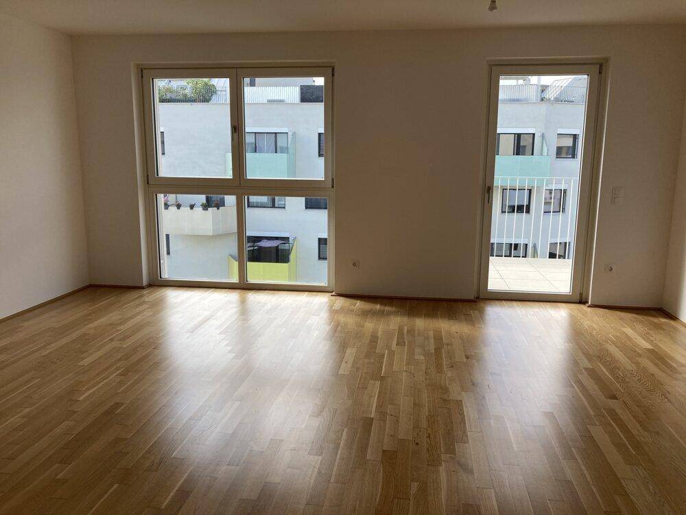 Heller Wohnraum mit Holzboden und großen Fenstern, die viel Tageslicht hereinlassen und einen Balkonzugang bieten.