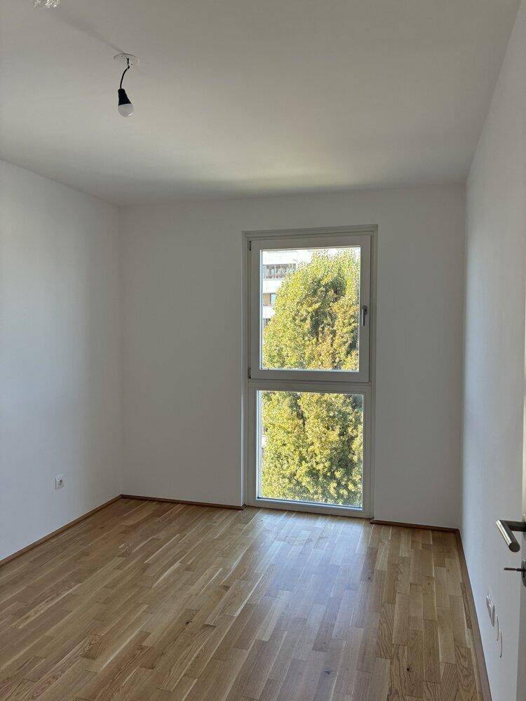 Helles Schlafzimmer mit Parkettboden und großem Fenster mit Blick auf einen Baum.