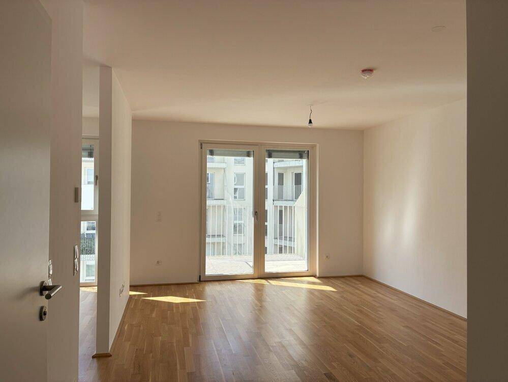 Unmöblierter Wohnraum mit Holzboden und Zugang zu einem Balkon, viel Tageslicht.