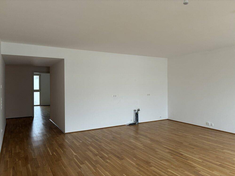 Heller, leerer Wohnbereich mit Parkettboden und offenem Übergang zu einem weiteren Raum.