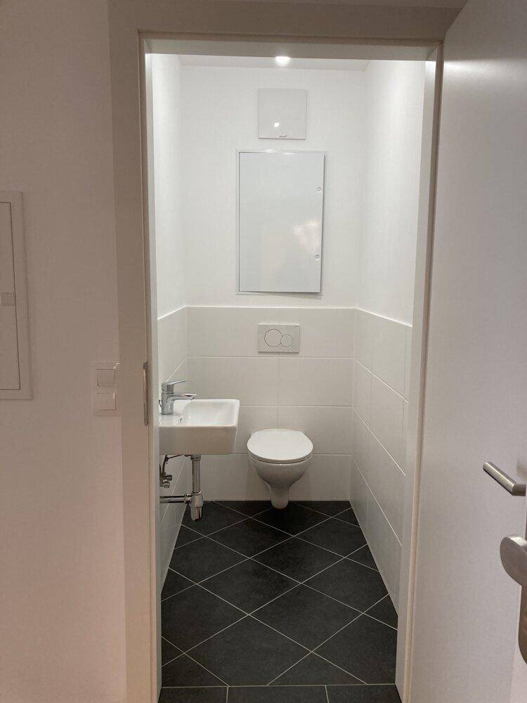 Separates Gäste-WC mit wandhängender Toilette, kleinem Waschbecken und dunklen Bodenfliesen.