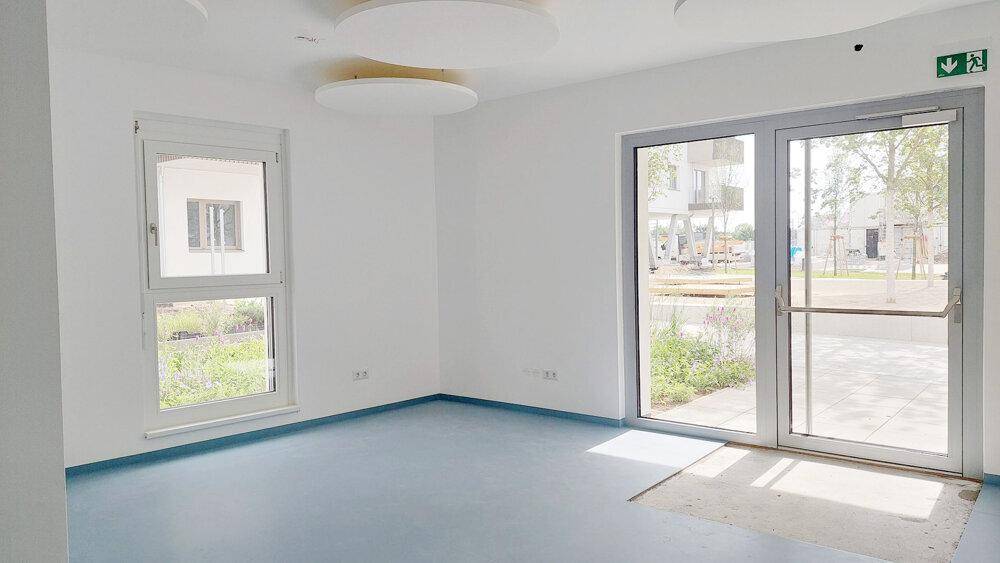 Heller, leerer Raum mit großen Fenstern, blauer Bodenfläche und direktem Zugang nach draußen.