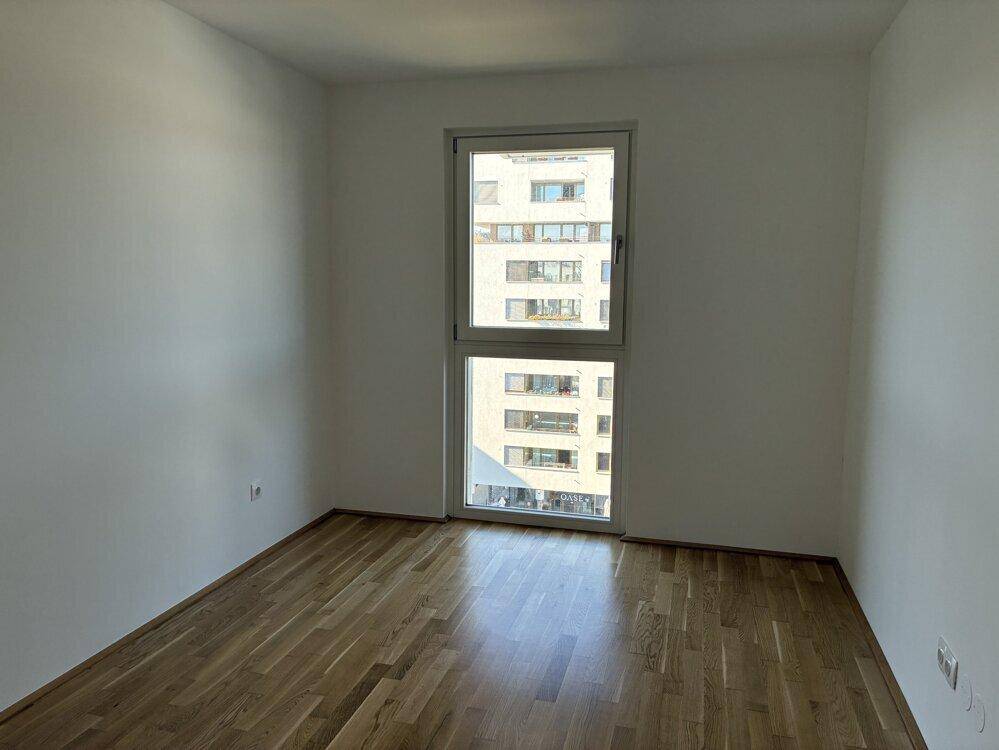 Helles Schlafzimmer mit Parkettboden und großem Fenster, das einen urbanen Ausblick bietet.