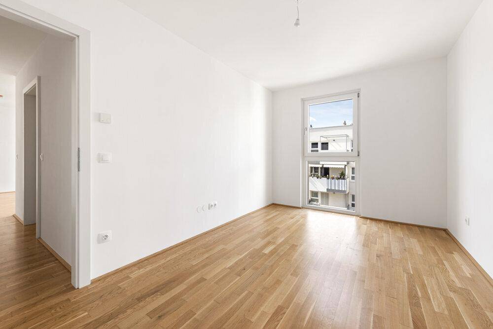 Geräumiges, helles Zimmer mit Holzboden und Fenster, das viel Tageslicht hereinlässt.