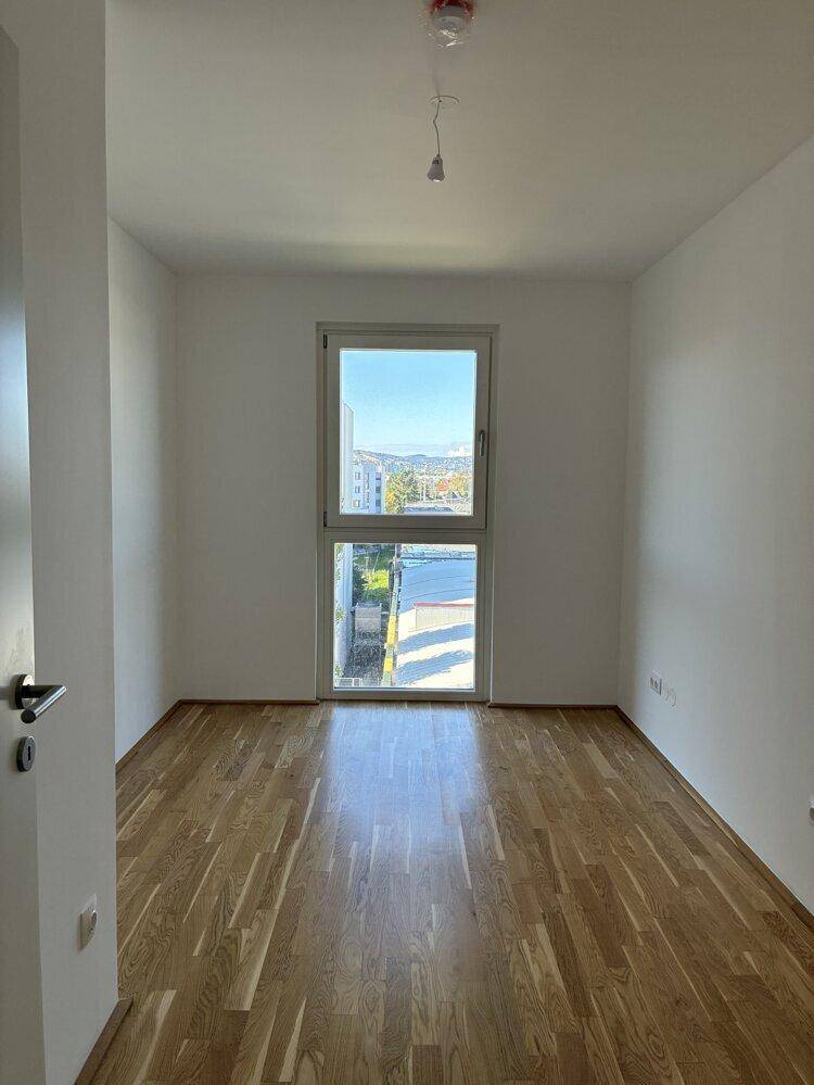 Helles Schlafzimmer mit Parkettboden und großem Fenster, das viel Tageslicht hereinlässt.