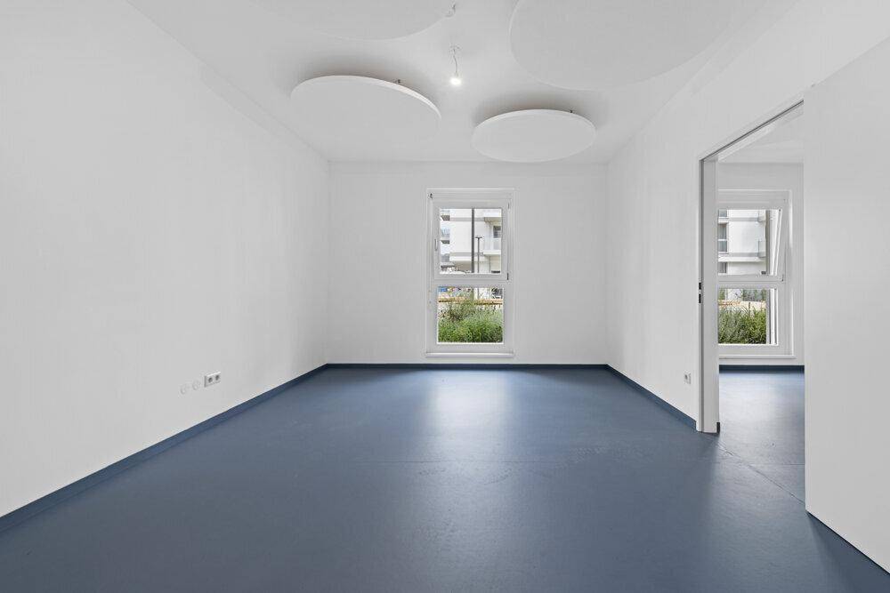 Heller, leerer Raum mit blauer Bodenfläche, weißen Wänden und einem Fenster mit Blick ins Grüne.