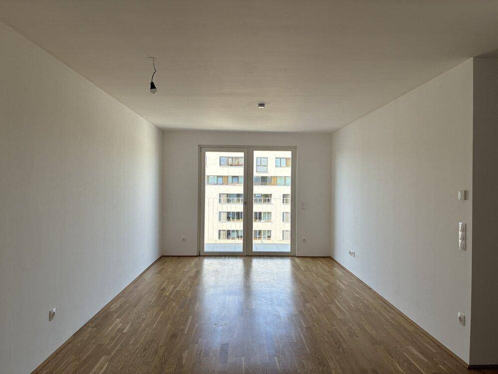 Heller, leerer Raum mit Holzboden und großen Fenstern, die viel Licht hereinlassen.