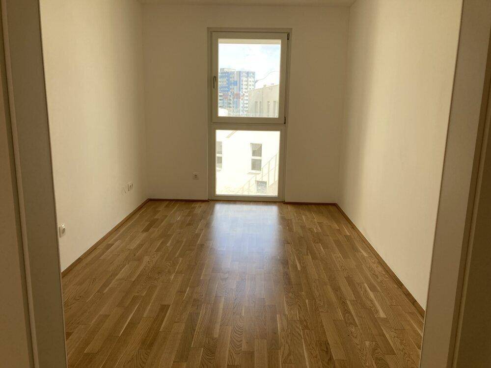 Leeres Schlafzimmer mit Parkettboden und einem großen Fenster, das einen Blick auf die umliegenden Gebäude bietet.