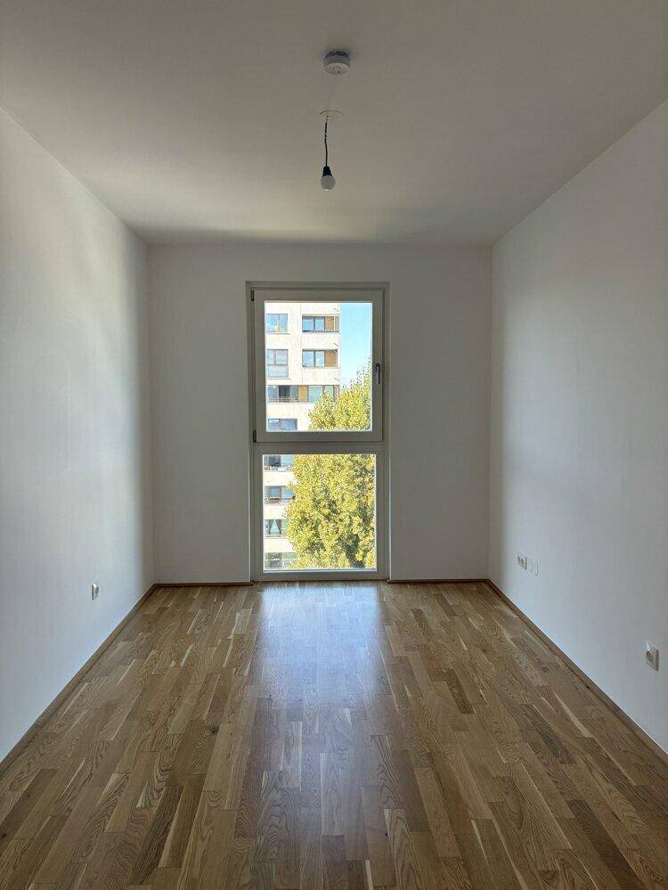 Helles Zimmer mit Holzboden und einem hohen Fenster, das viel Tageslicht bietet.