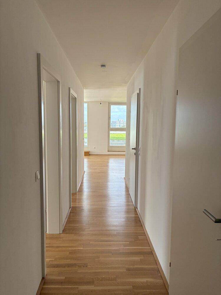 Heller Flur mit Holzboden und weißen Wänden, der zu einem lichtdurchfluteten Raum führt.