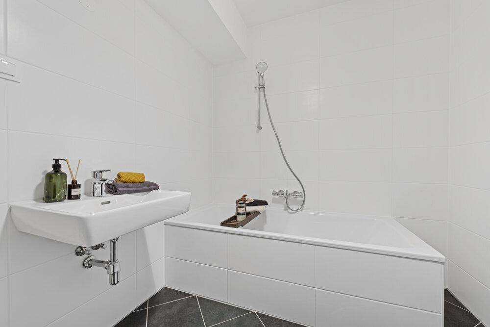 Helles Badezimmer mit weißen Fliesen, dunklem Boden, Badewanne und modernem Waschbecken.