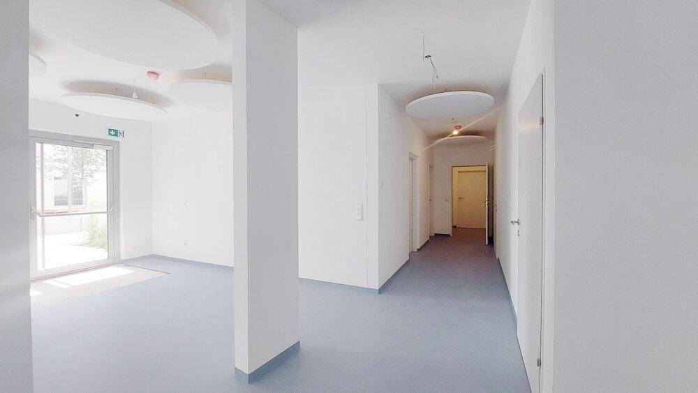Heller Flur mit Zugang nach draußen, weißen Wänden, blauem Boden und modernen Deckenelementen.