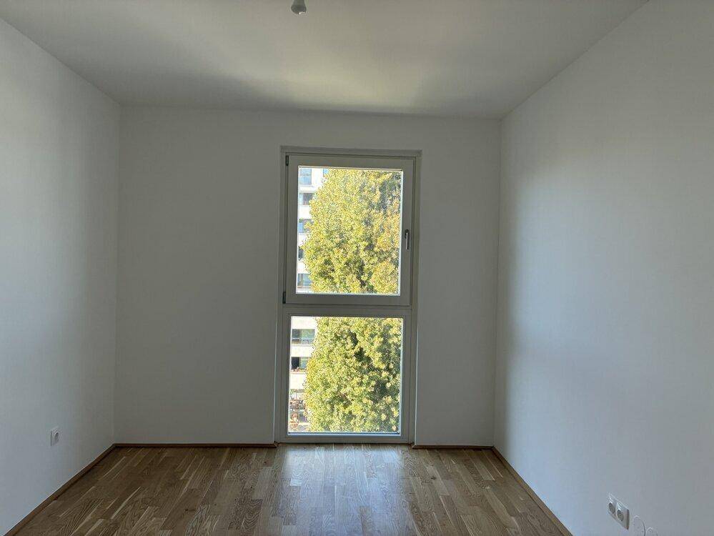 Helles Zimmer mit Parkettboden und großem Fenster mit Blick ins Grüne.
