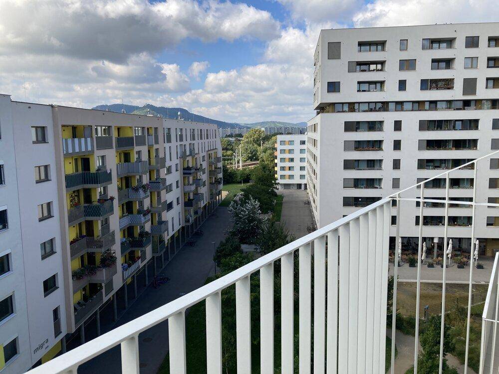 Ausblick von einem Balkon auf moderne Wohngebäude und eine grüne Umgebung mit Bergen im Hintergrund.