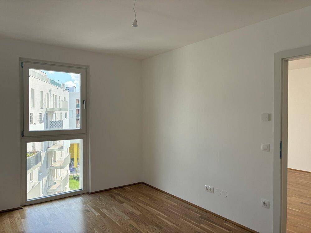 Helles Schlafzimmer mit Parkettboden und großem Fenster, das einen Blick ins Grüne bietet.