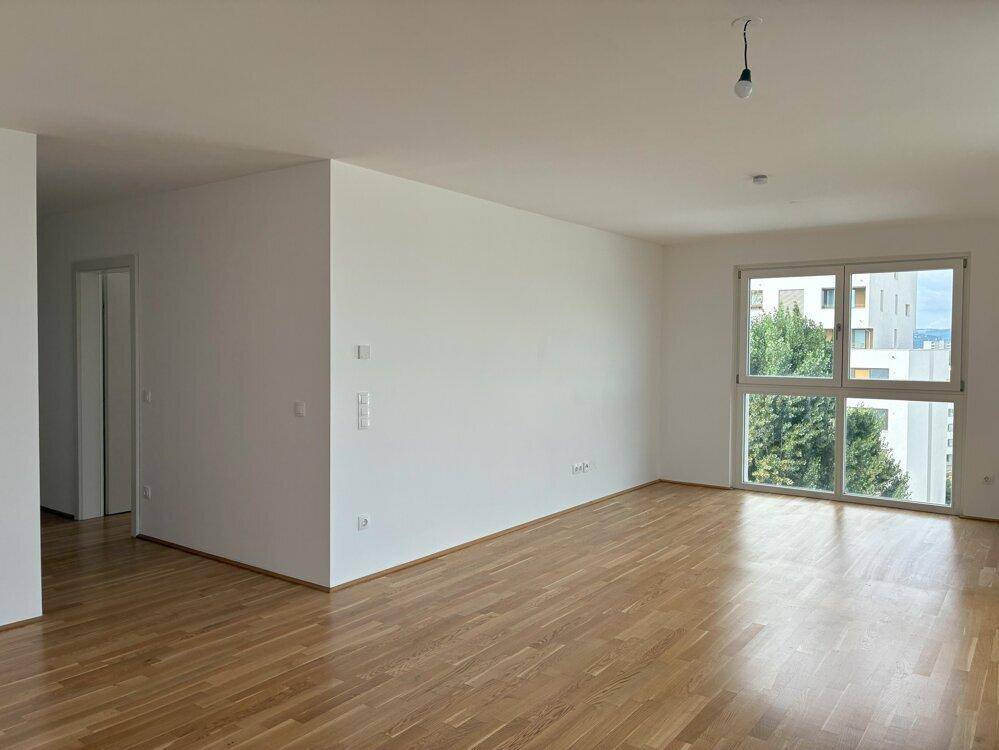 Geräumiger, leerer Raum mit Holzboden und einem großen Fenster, das viel Licht hereinlässt.