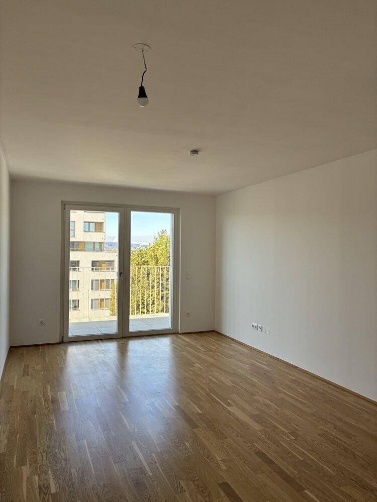 Helles, leeres Zimmer mit Holzboden und Zugang zu einem Balkon.