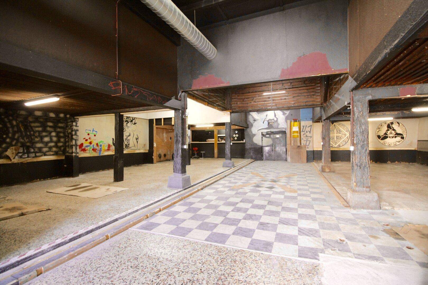 Großer Innenraum mit kariertem Fliesenboden, Holzbalken und Graffiti an den Wänden.