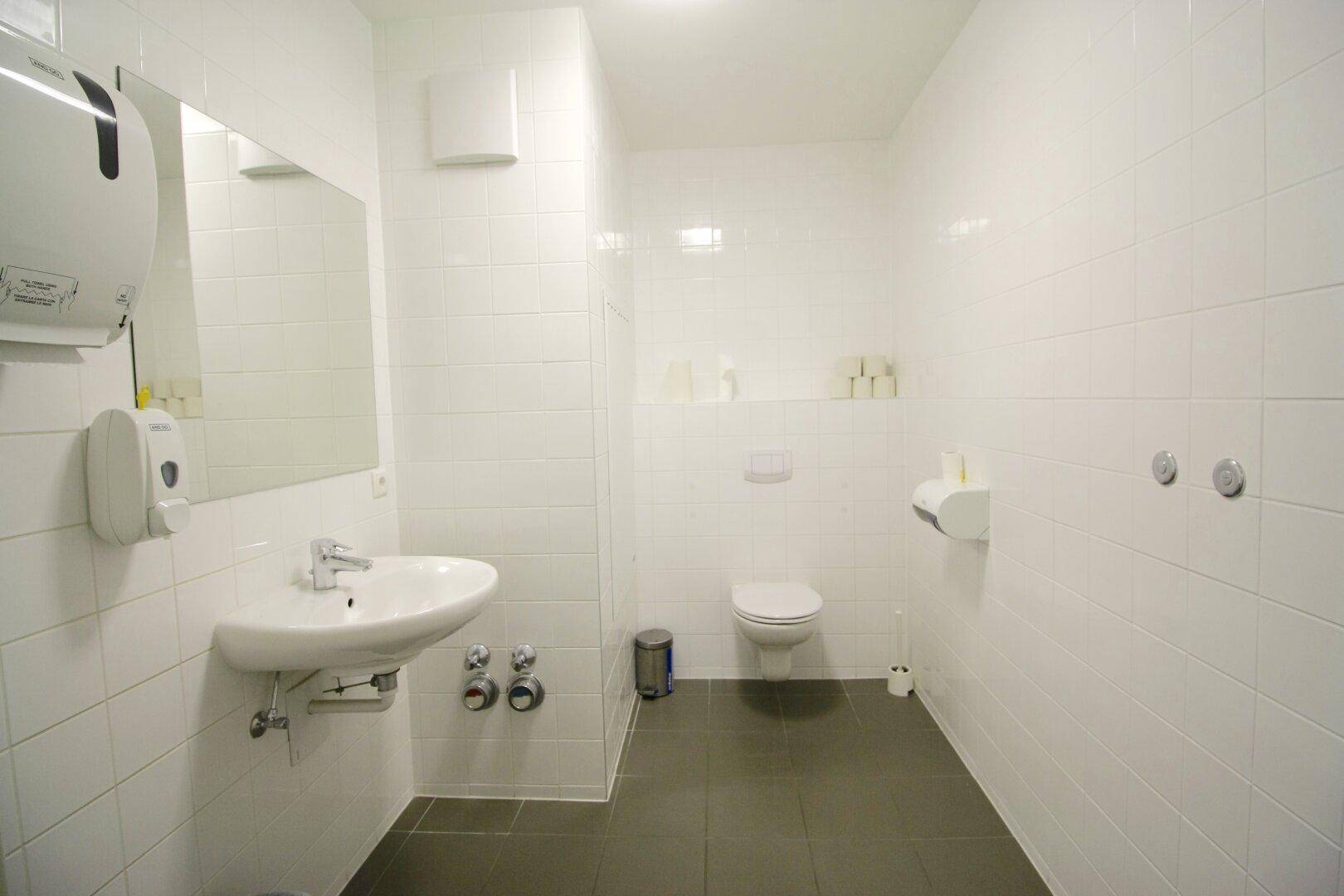 Helle und saubere Toilette mit weißen Fliesen, dunklem Boden und Waschbecken.