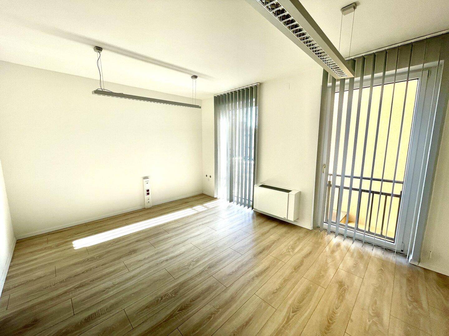 Geräumiger Bürobereich mit viel Tageslicht, vertikalen Jalousien und hellem Laminatboden.