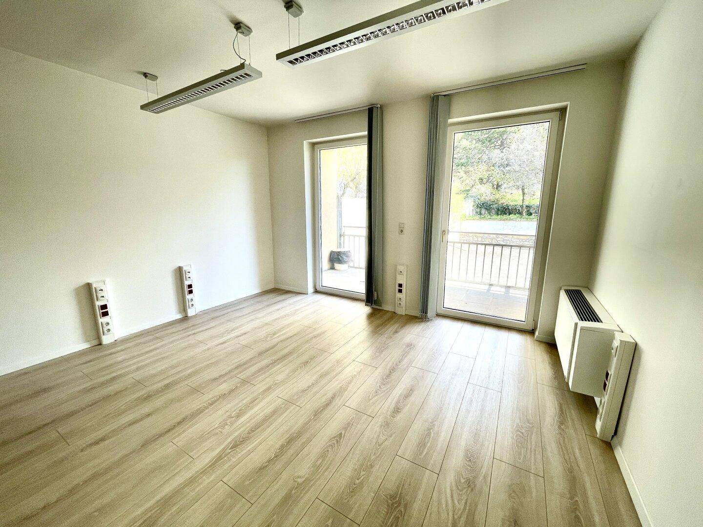 Leerer Büroraum mit zwei Fenstertüren, die auf einen Balkon führen, und hellem Laminatboden.