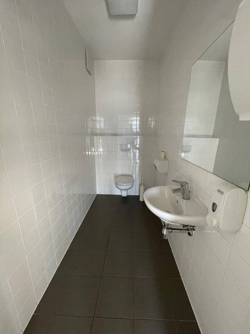 Einfache Toilette mit weißen Fliesen, dunklem Boden und Waschbecken mit Spiegel.