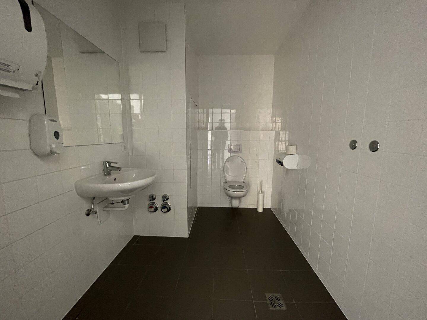 Saubere Toilette mit weißen Wandfliesen, dunklem Boden und Waschbecken mit Spiegel.
