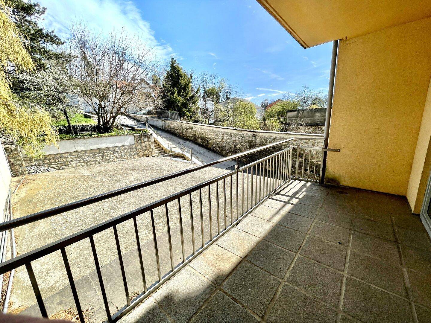 Balkon mit Fliesenboden und Metallgeländer, bietet Blick auf eine Auffahrt und Bäume.