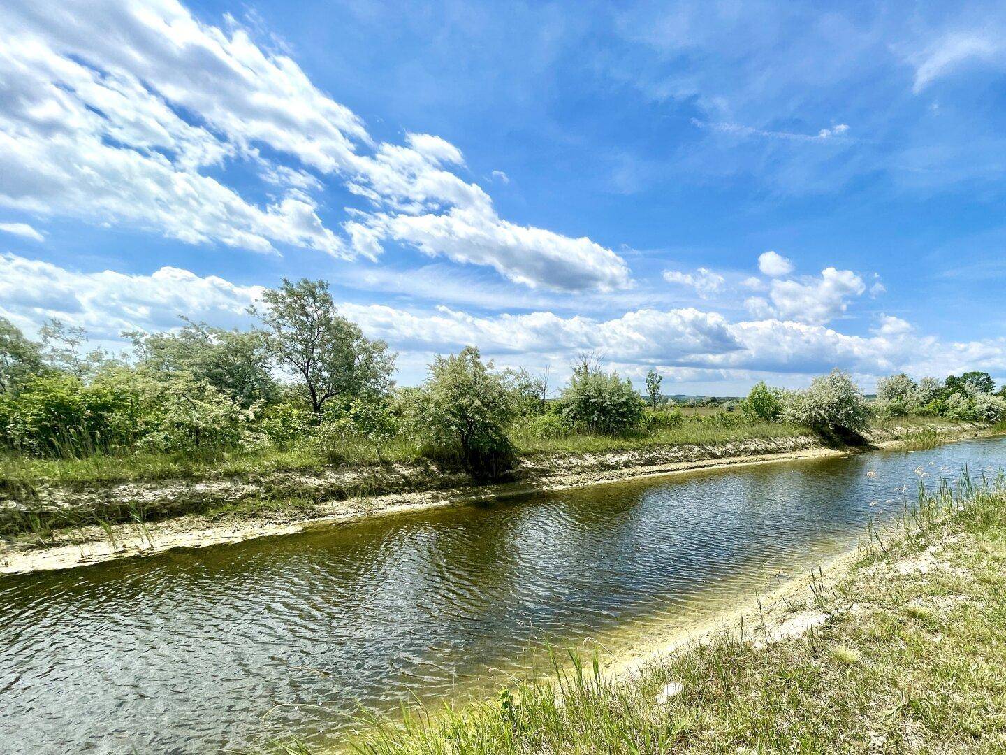 Ruhiger Wasserlauf mit grünen Ufern und Bäumen unter einem weiten blauen Himmel.
