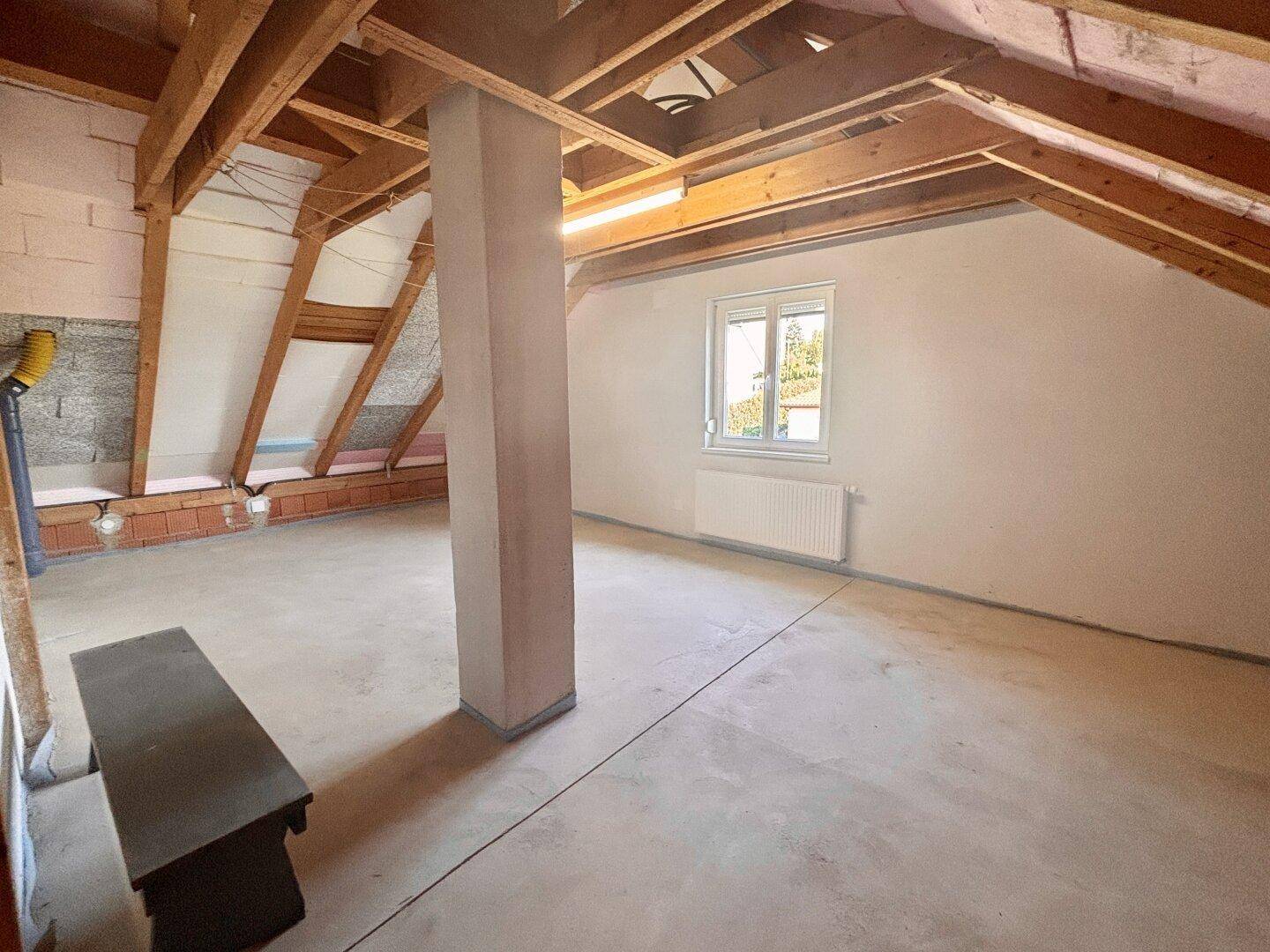Heller Dachbodenraum mit Fenster, sichtbaren Holzbalken und Betonboden, bereit zum Ausbau.