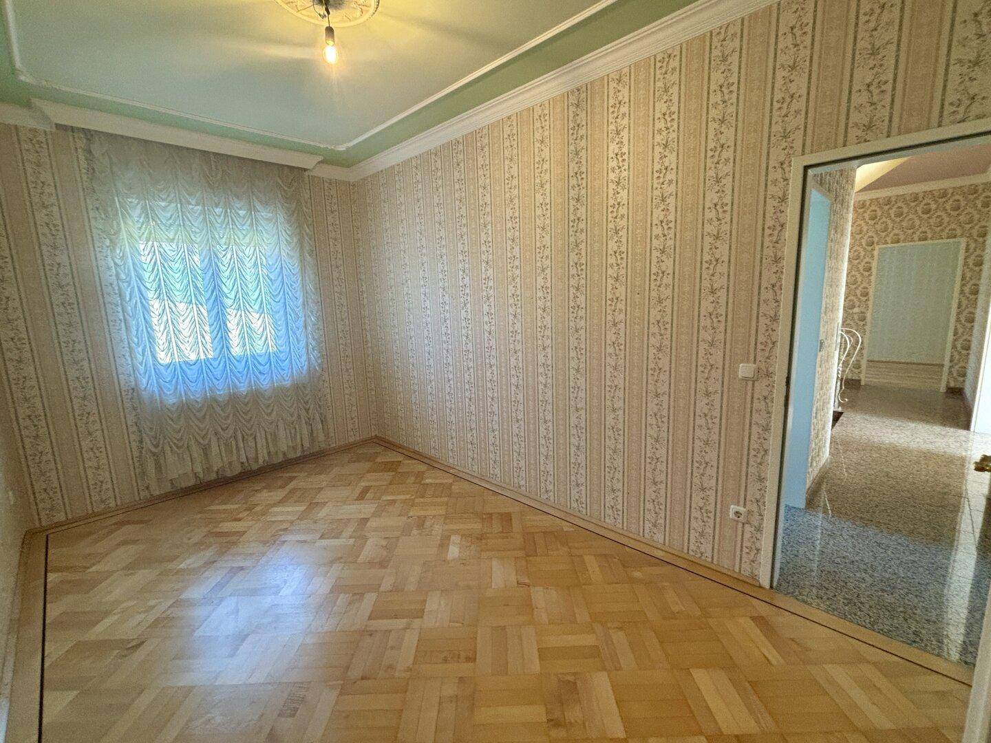 Schlafzimmer mit Parkettboden, gemusterter Tapete und Fenster, das Tageslicht hereinlässt.