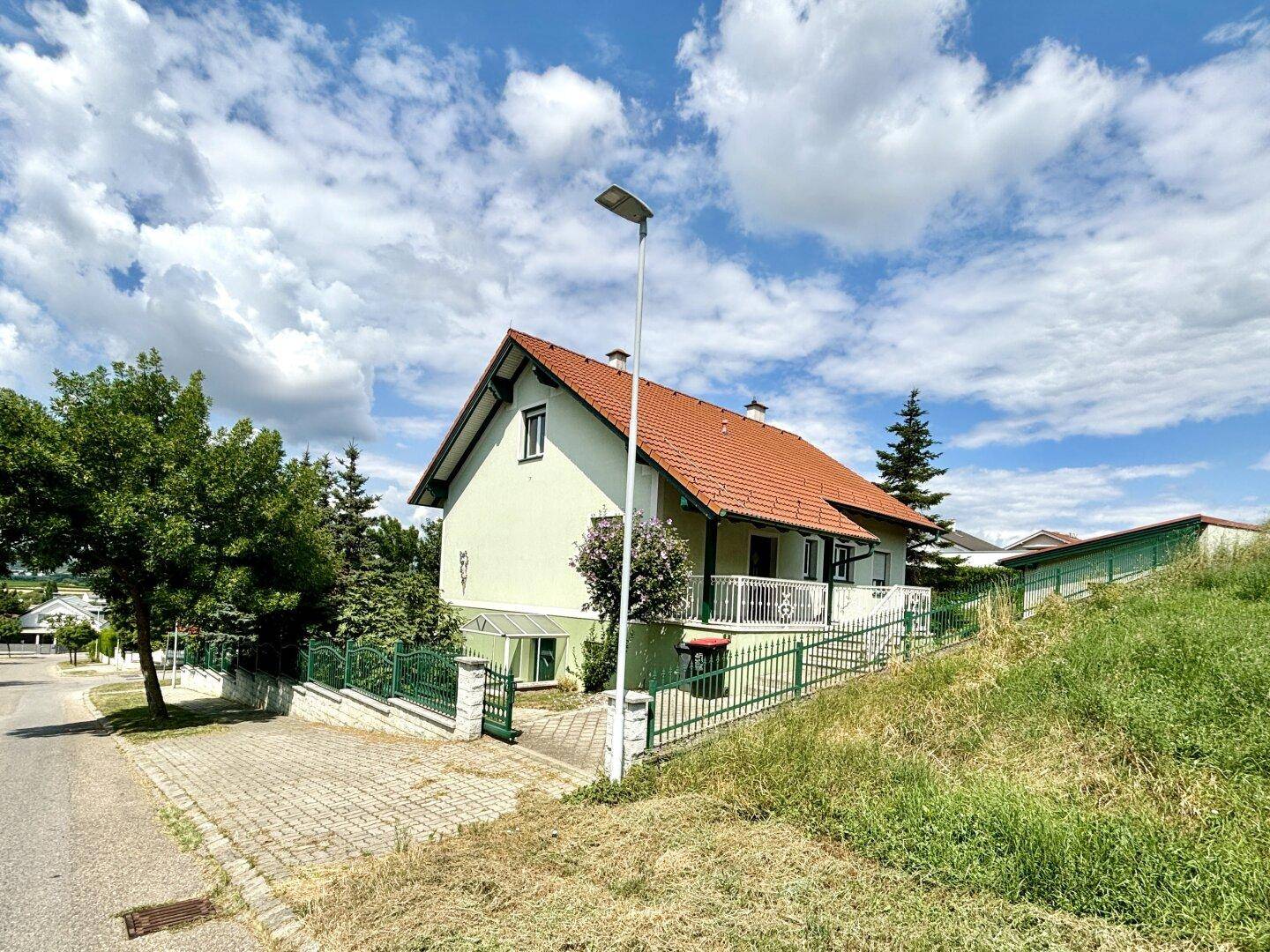 Freistehendes Einfamilienhaus mit rotem Ziegeldach und grünem Zaun in ländlicher Umgebung.