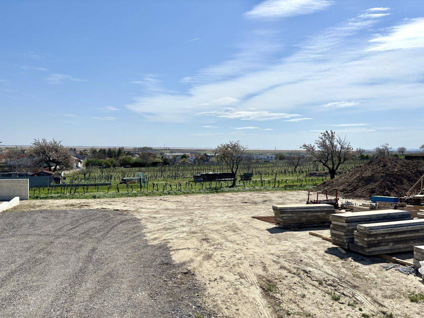 Baugrundstück mit Schotter und Baumaterialien, umgeben von Weinbergen und einem Dorf in der Ferne.
