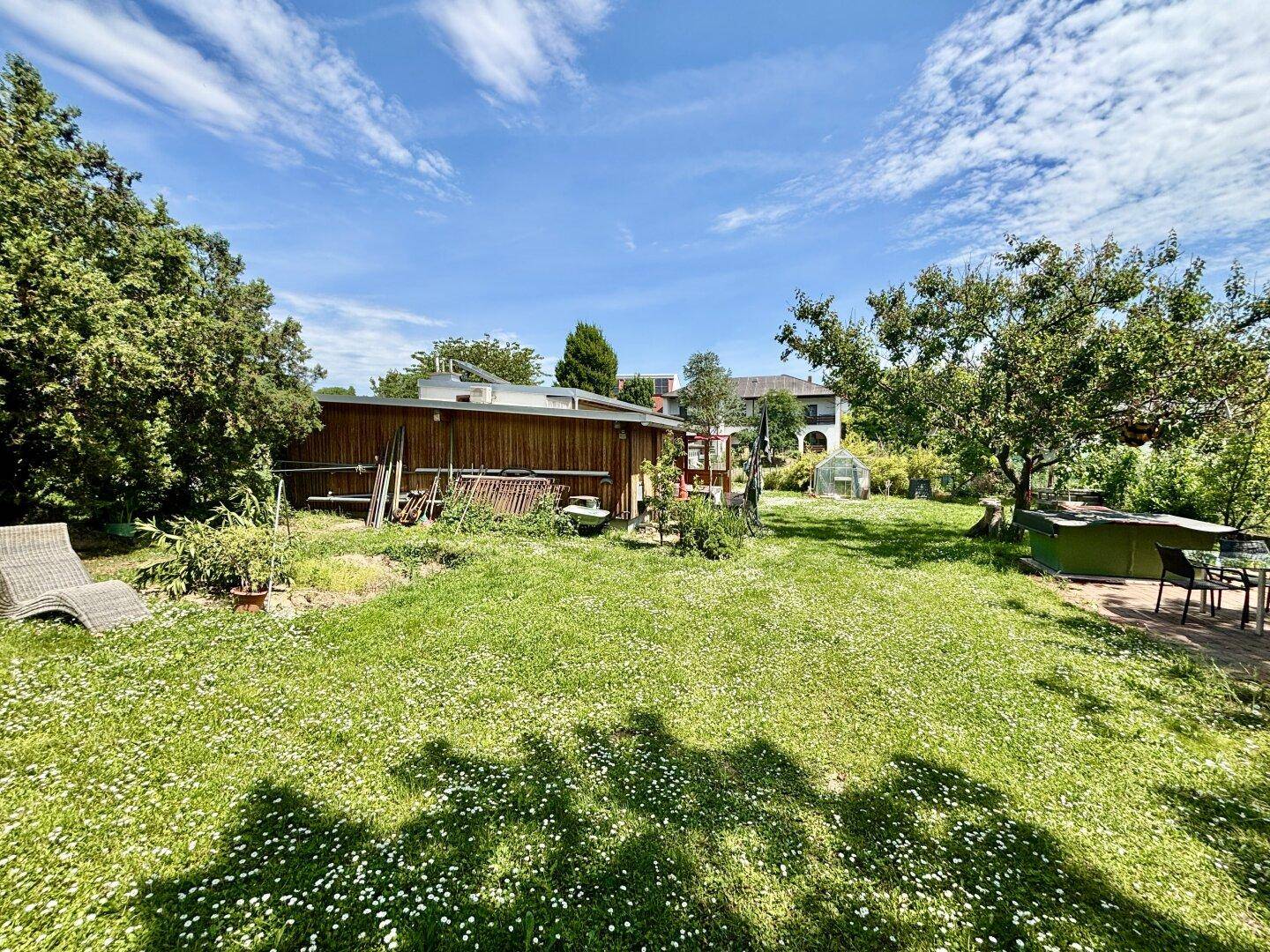 Großer Garten mit Rasenfläche, Liegestuhl und Blick auf das Wohnhaus mit Gewächshaus.