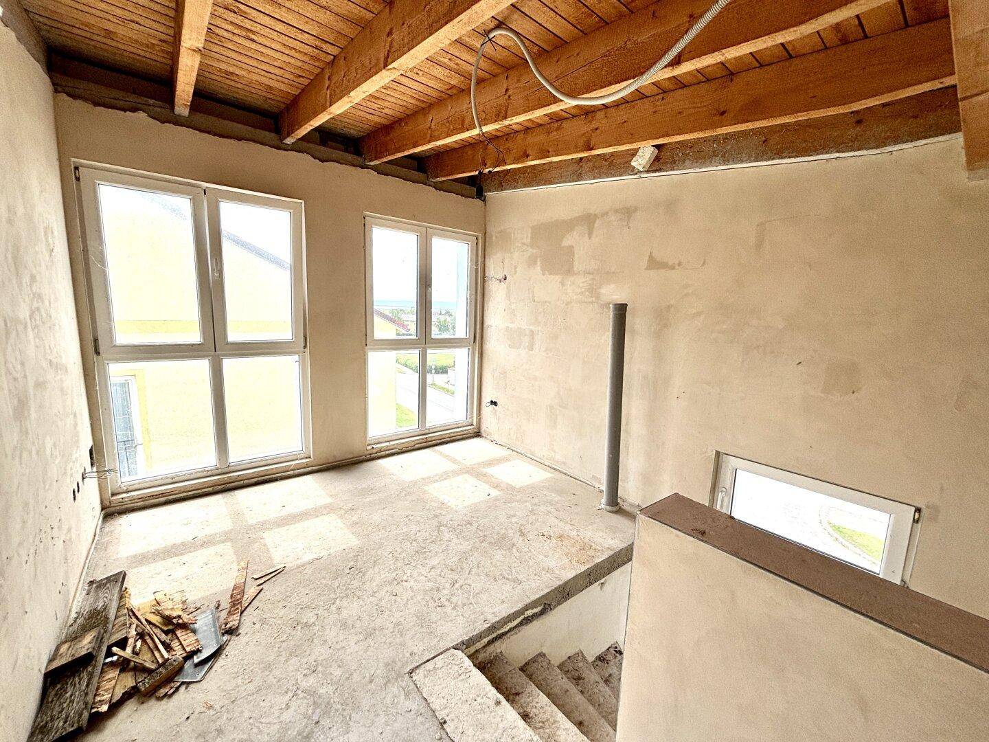 Unfertiger Innenraum mit sichtbaren Holzbalken an der Decke und großen Fenstern.