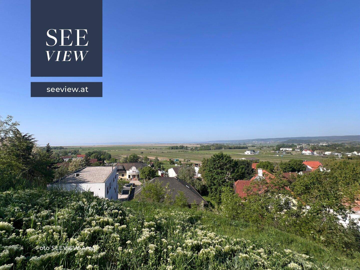 Panoramablick über eine grüne Landschaft mit kleinen Häusern und einem Seehorizont unter klarem, blauem Himmel.