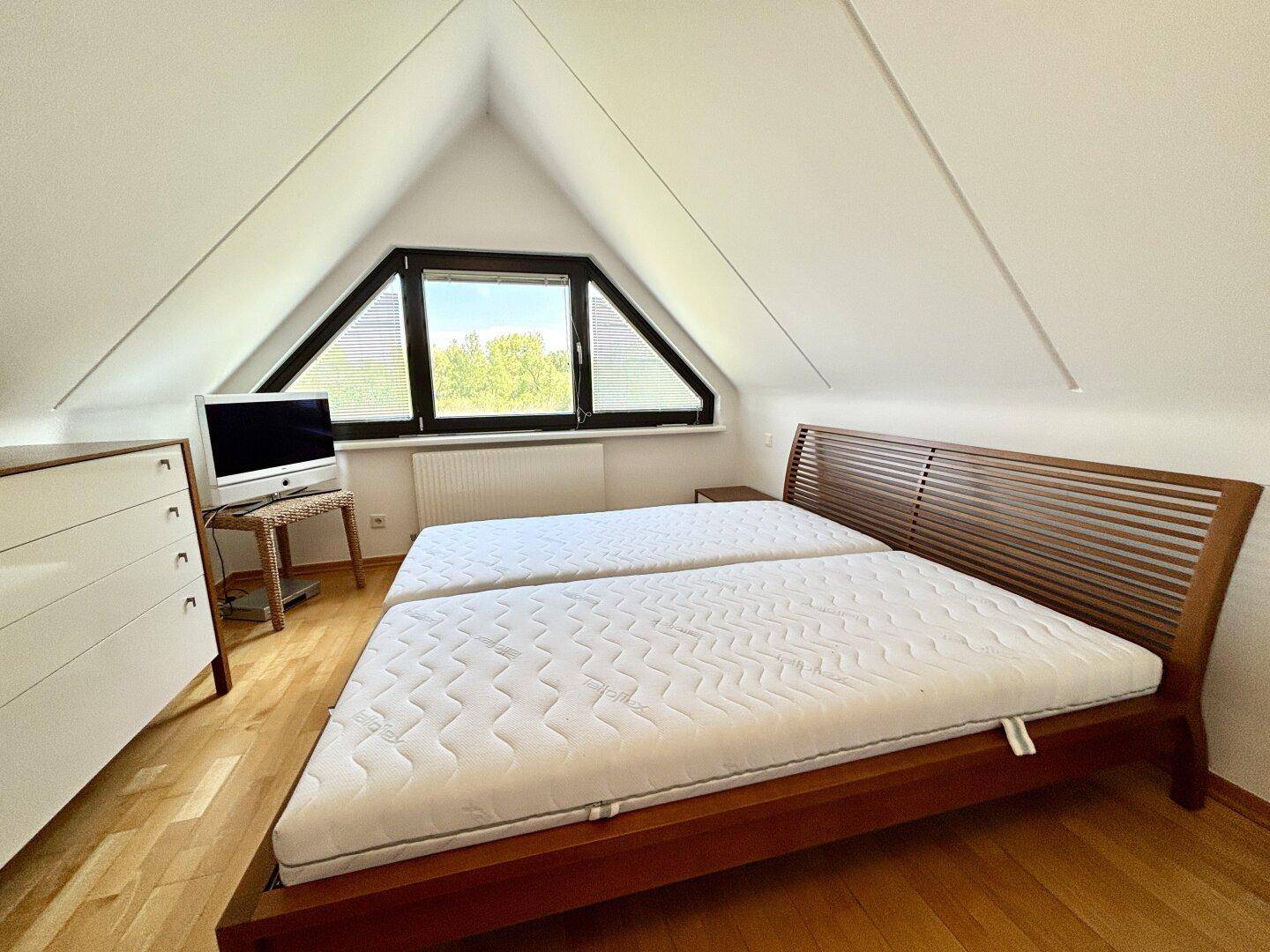 Gemütliches Schlafzimmer im Dachgeschoss mit Holzboden, Doppelbett und dreieckigem Fenster.